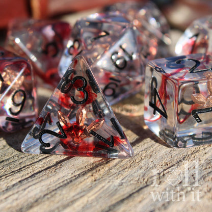 Clear dice with black numbers and rose gold miniature daggers and knives inside on a wooden surface, featuring the brand 'Roll with It'.