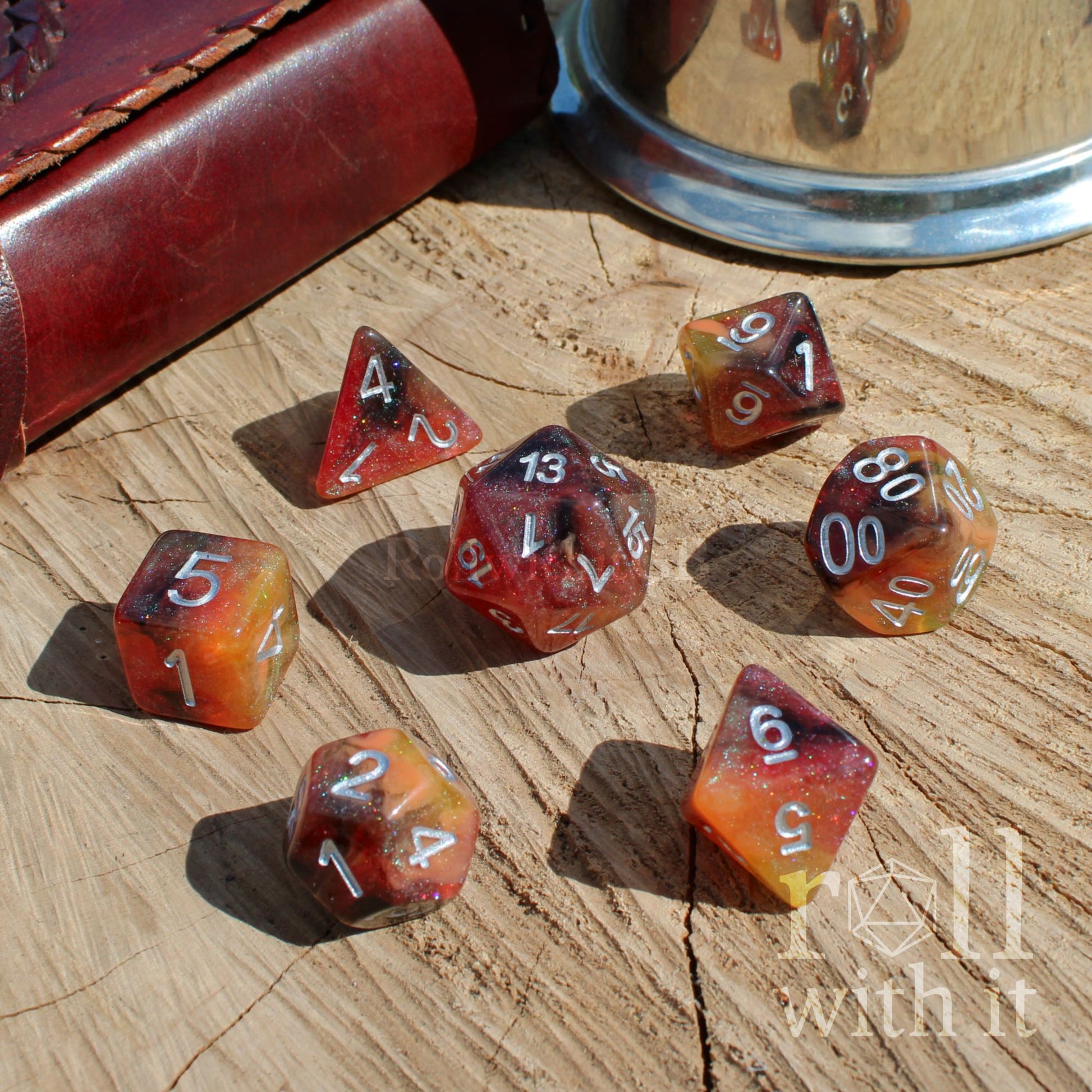 Set of polyhedral burnt orange dark glittery mixed colour dice on a wooden surface with a leather-bound book and metal tray in the background.
