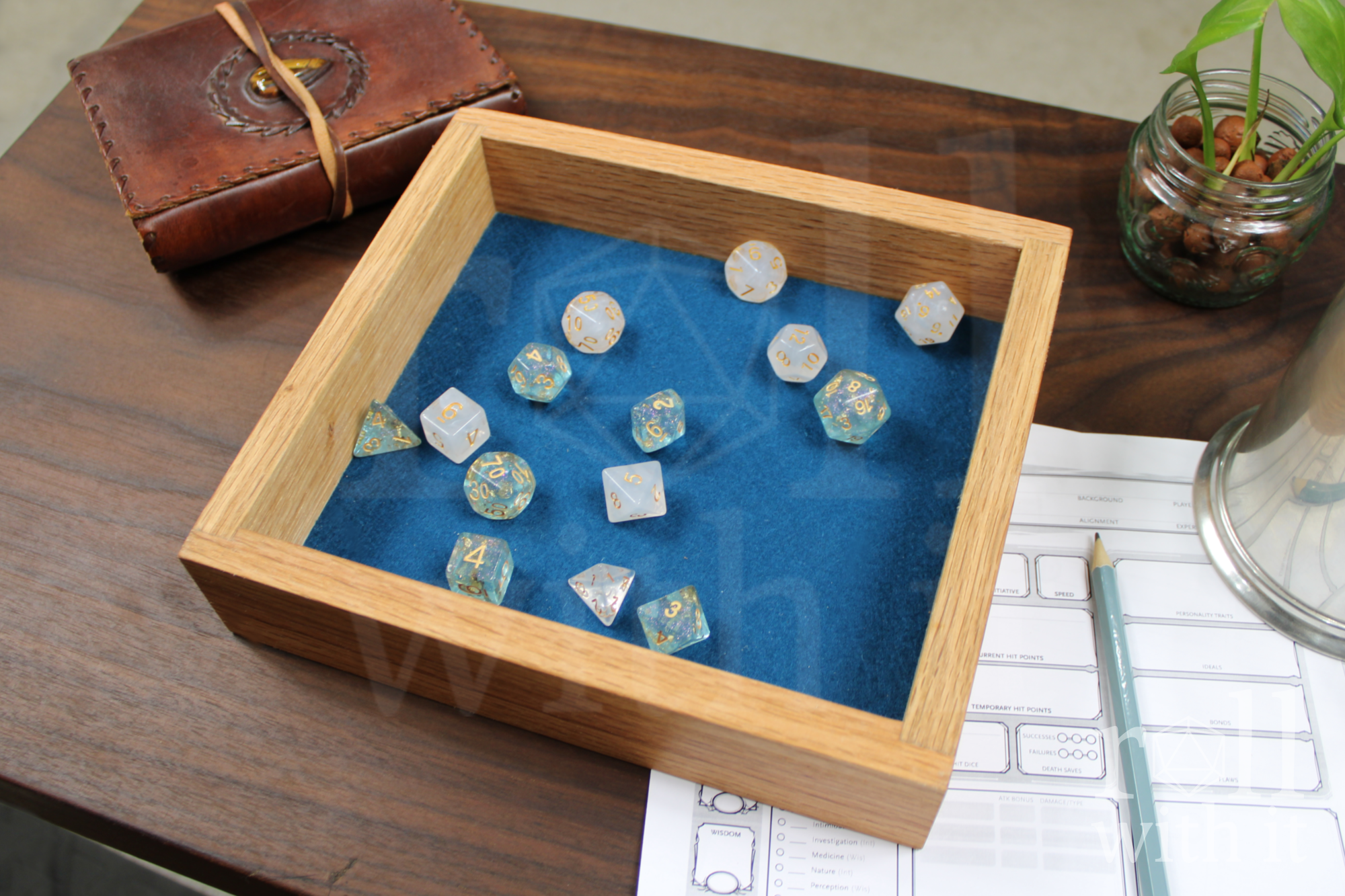 Red oak dice tray with ice blue and white polyhedral dice sit on a navy blue felt interior base on a wooden table, featuring 'roll with it' logo. A character sheet and other tabletop accessories are in the background.