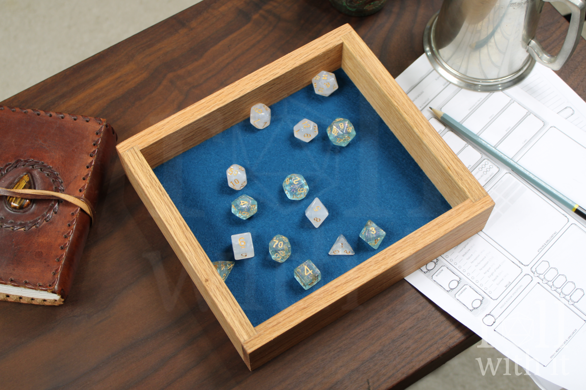 Wooden red oak dice tray with a set of ice blue and white roleplaying dice sit on a navy blue felt interior on a wooden table, featuring 'roll with it' logo. Other tabletop accessories are in the background.