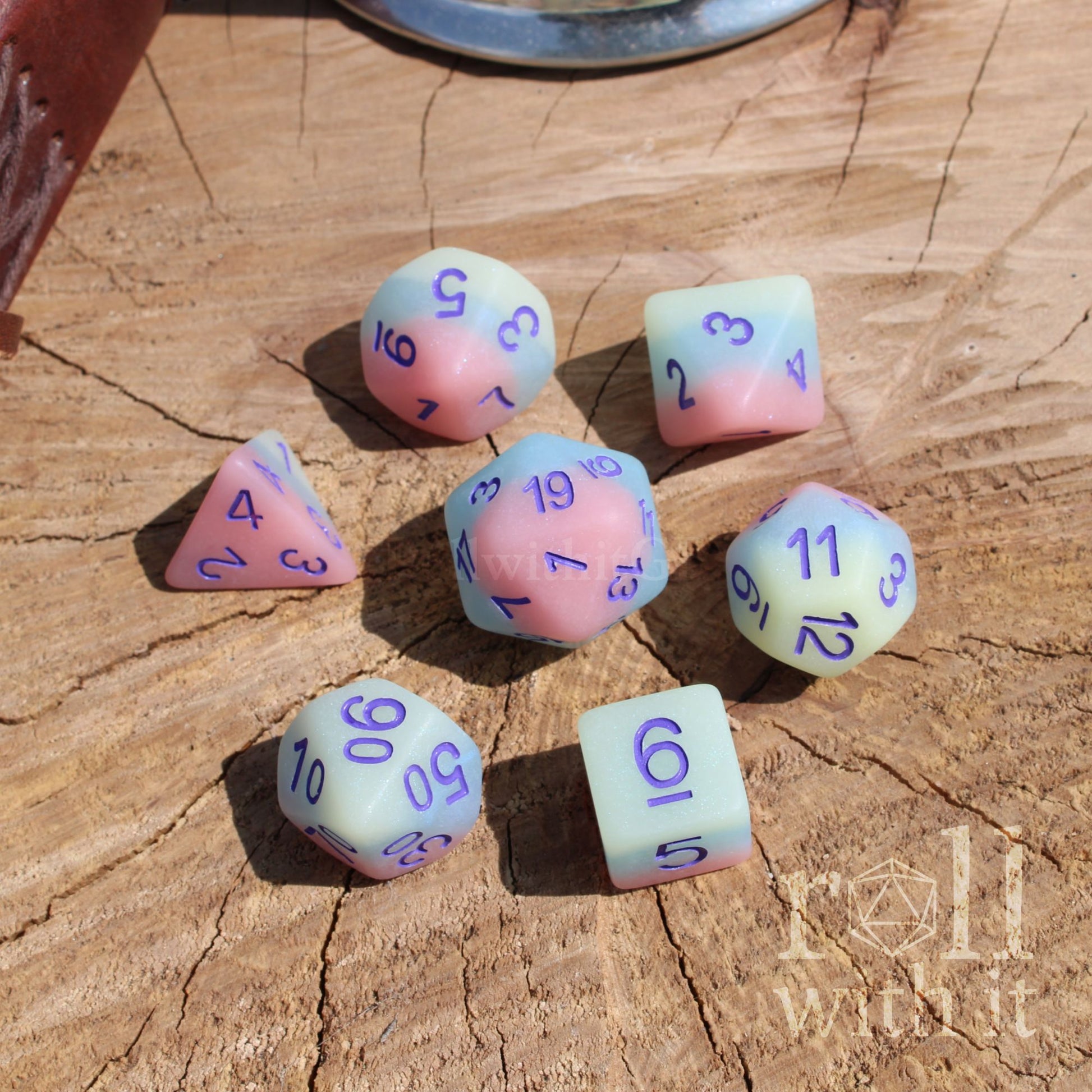 Set of pastel-colored pale blue pink yellow polyhedral dice with purple numbers on a wooden surface, featuring 'roll with it' branding.