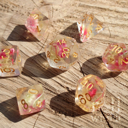 7 translucent polyhedral D&D dice with pink and gold flowers and golden numbers on a wooden surface