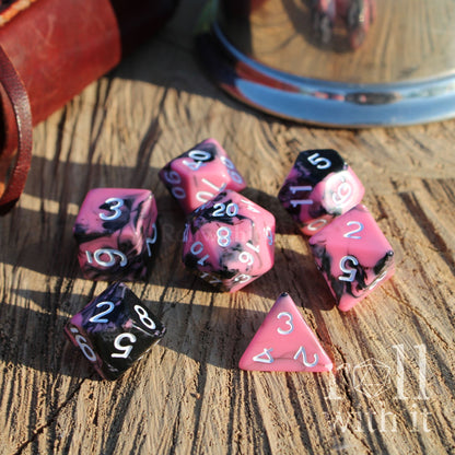 Set of pink and black mixed polyhedral dice on a wooden surface with a leather-bound book and glass in the background.