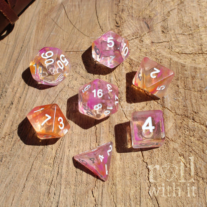 Set of pink and orange clear girly polyhedral dice on a wooden surface with 'roll with it' branding.