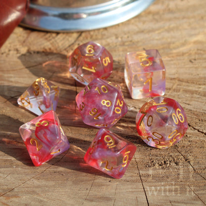 Set of 7 pink and clear polyhedral TTRPG dice with gold numbers on a wooden surface.
