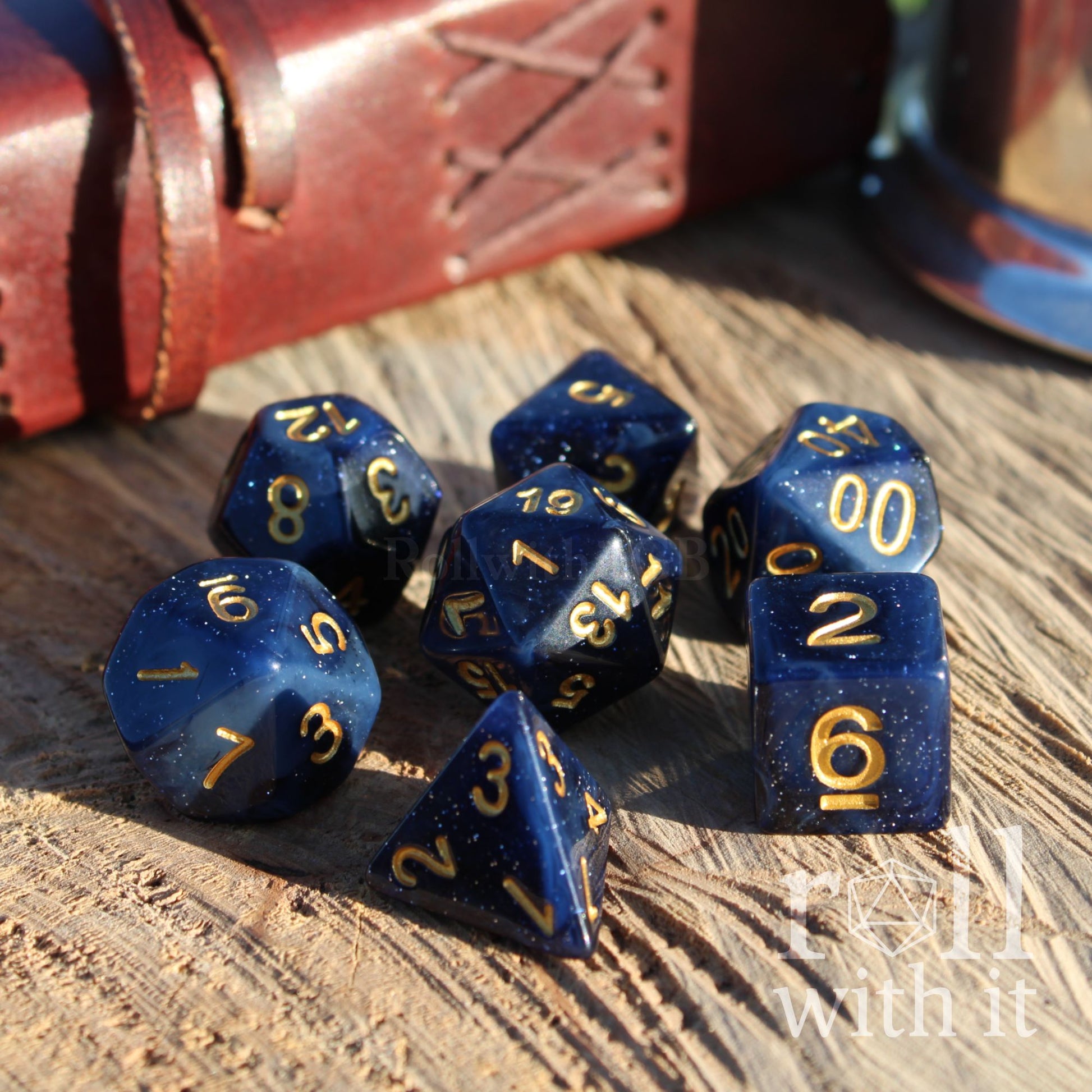 Deep dark blue polyhedral dice set with gold numbers on a wooden surface, with a leather-bound book in the background.