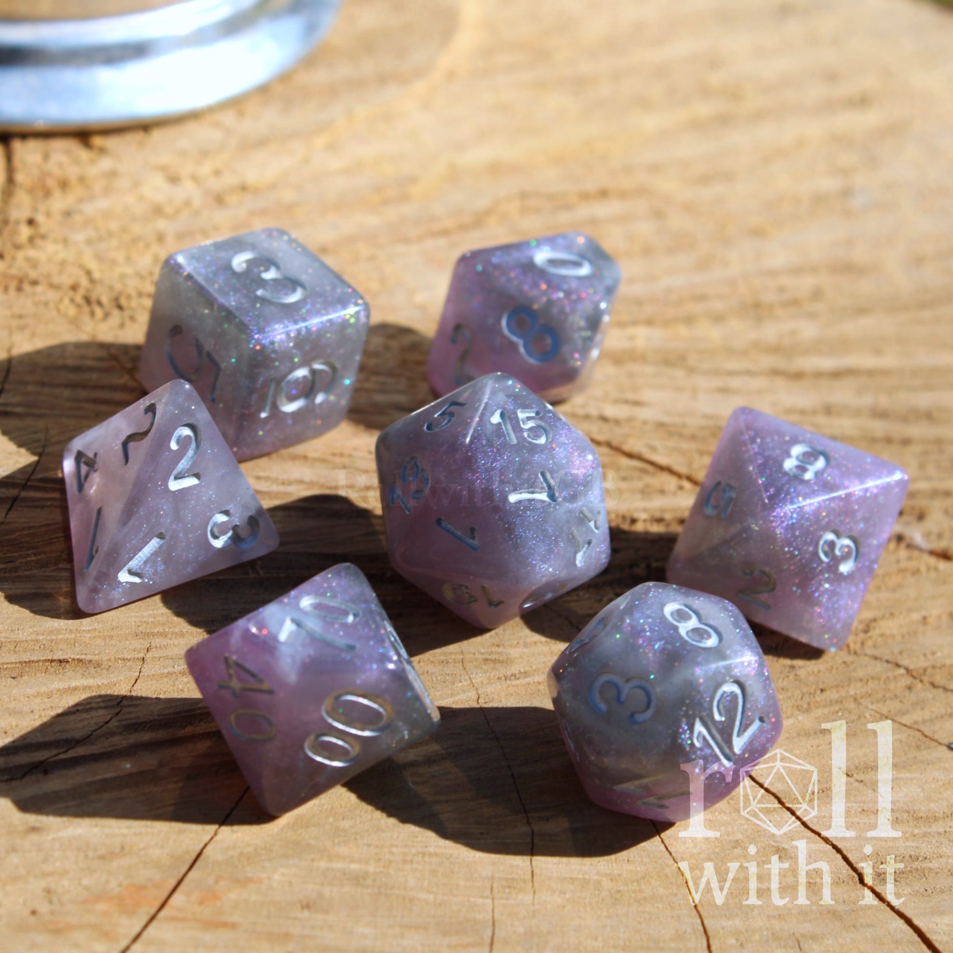 Set of purple silver galaxy star glitter D&D polyhedral dice on a wooden surface with 'Roll with It' logo.