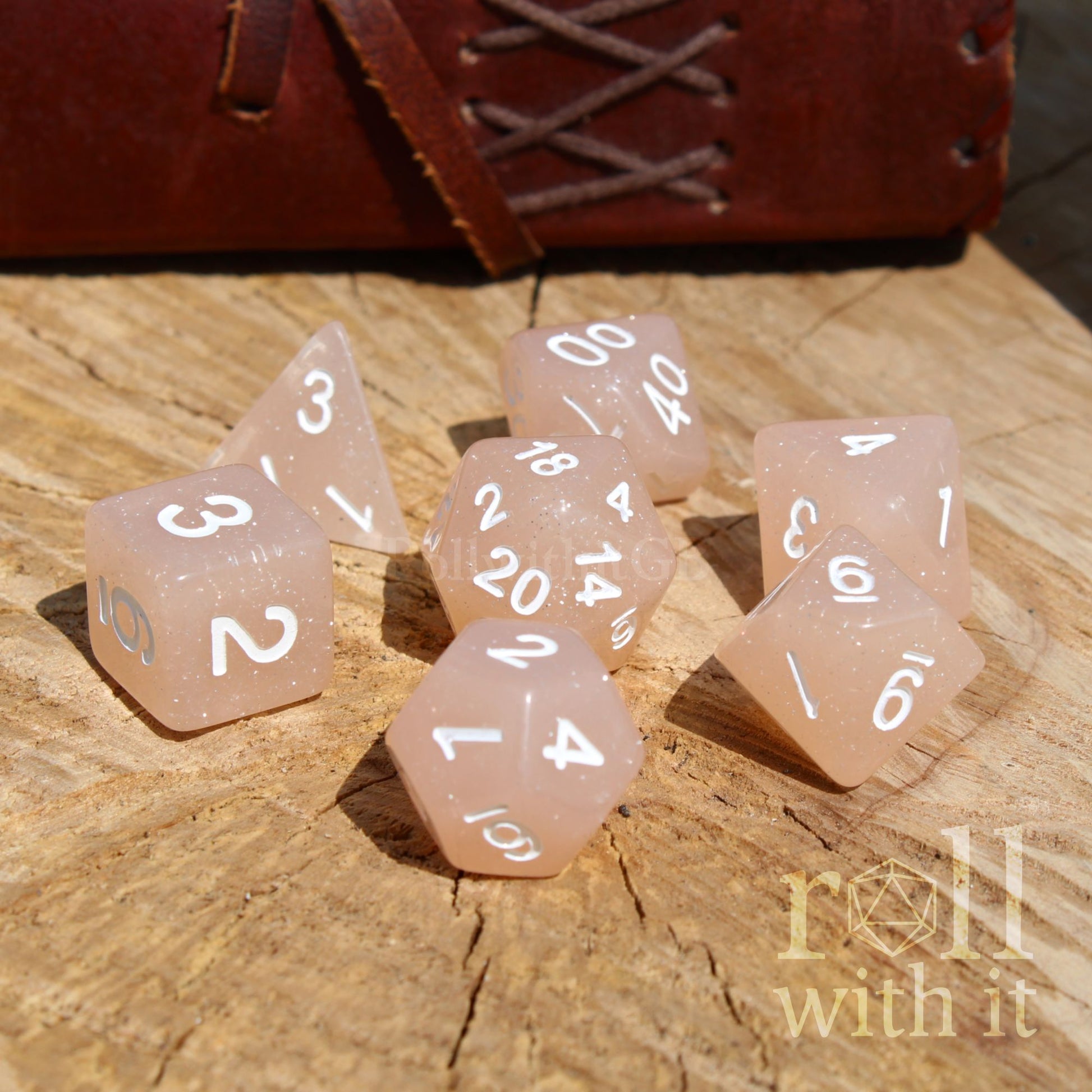 Set of rose coloured dice with white numbers on a wooden surface, featuring 'roll with it' branding.