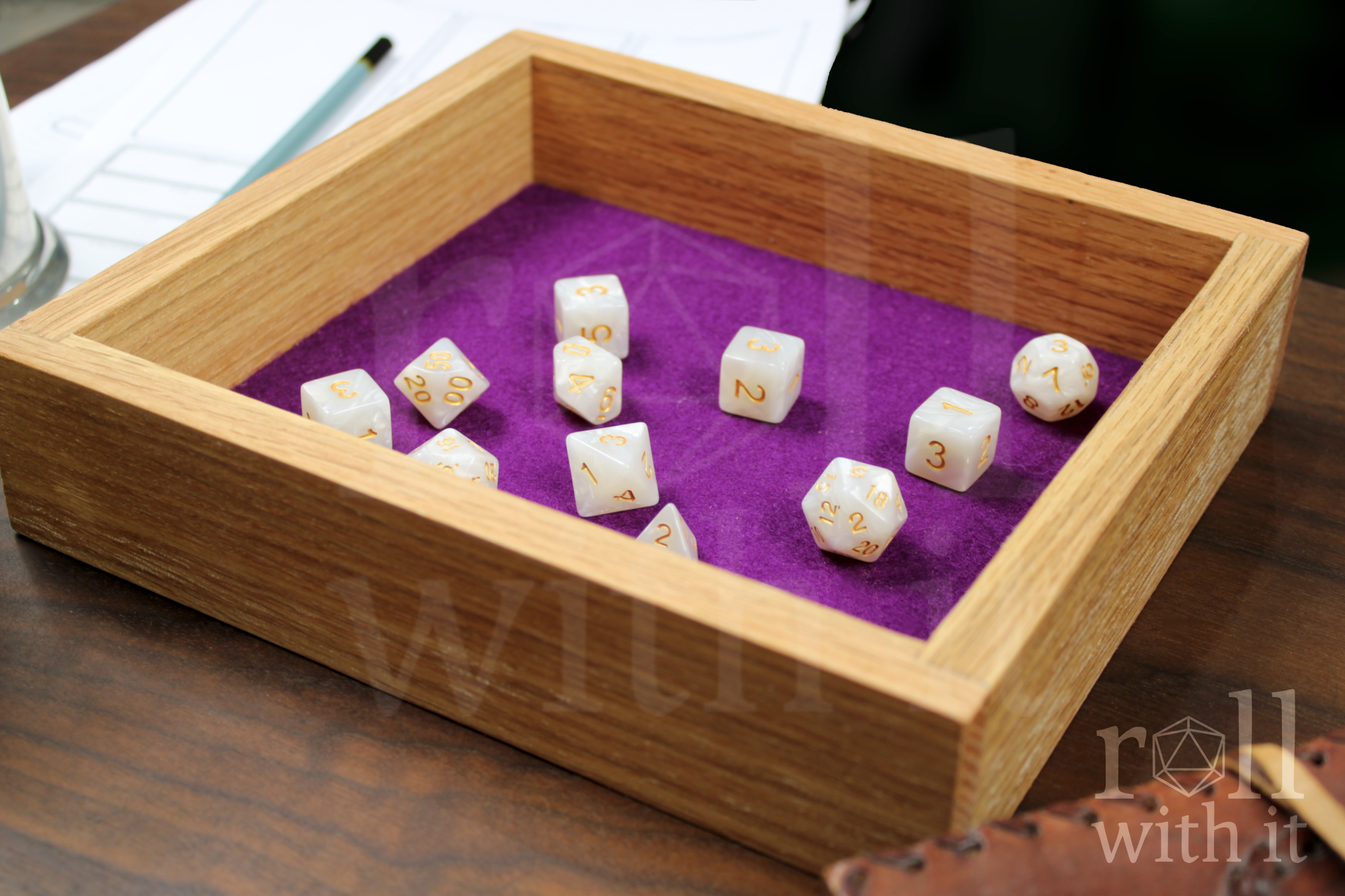 Wooden red oak dice tray with tyrian purple felt interior on a wooden table, featuring 'roll with it' logo. White pearl dice are inside the box. Other tabletop accessories are in the background.