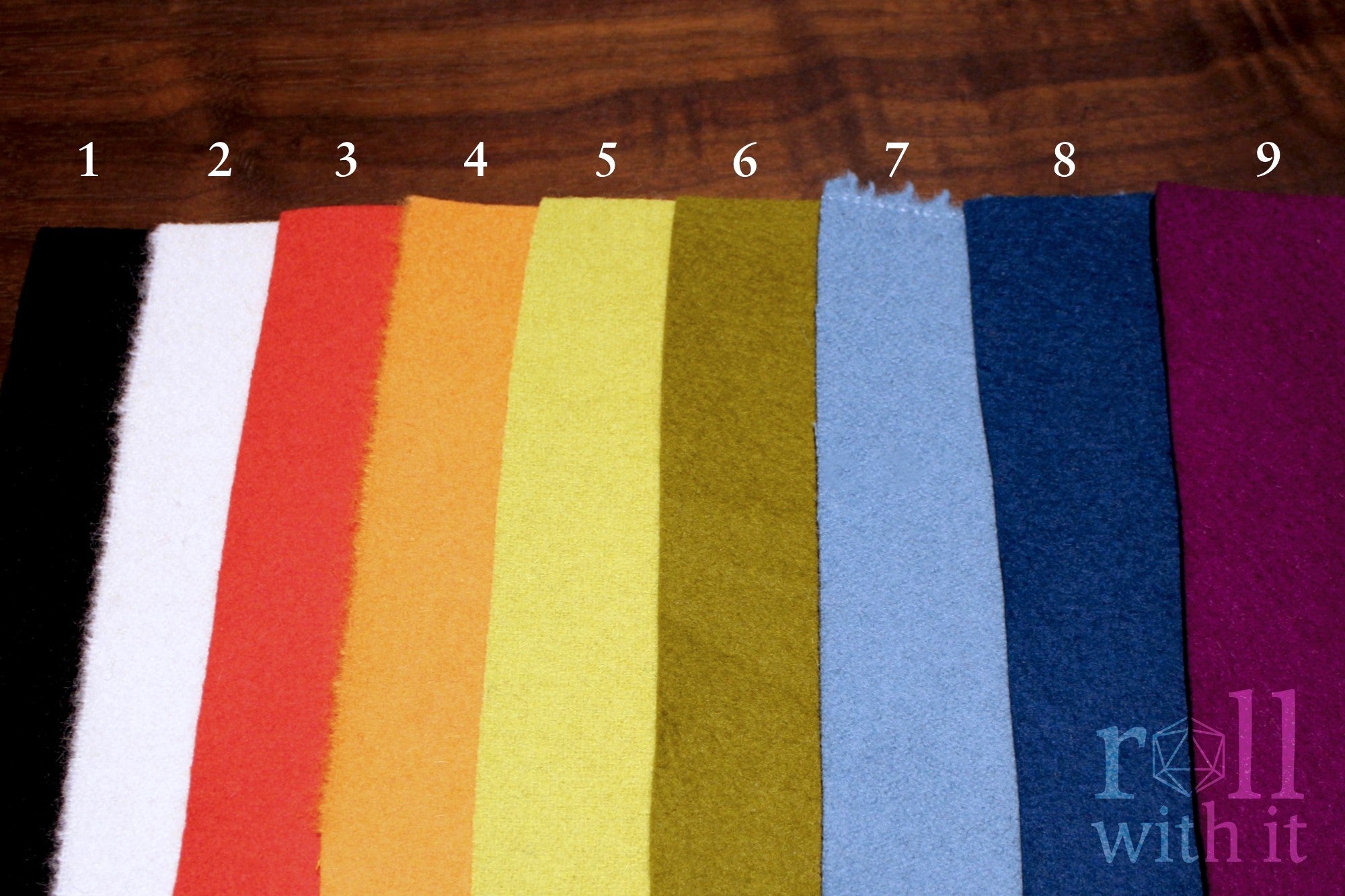 Nine wool felt fabric swatches in classic black, cream white, scarlet red, tangerine orange, chartreuse yellow, khaki green, robin egg blue, navy blue, and aubergine purple on a wooden surface with 'roll with it' branding in the corner of the image.