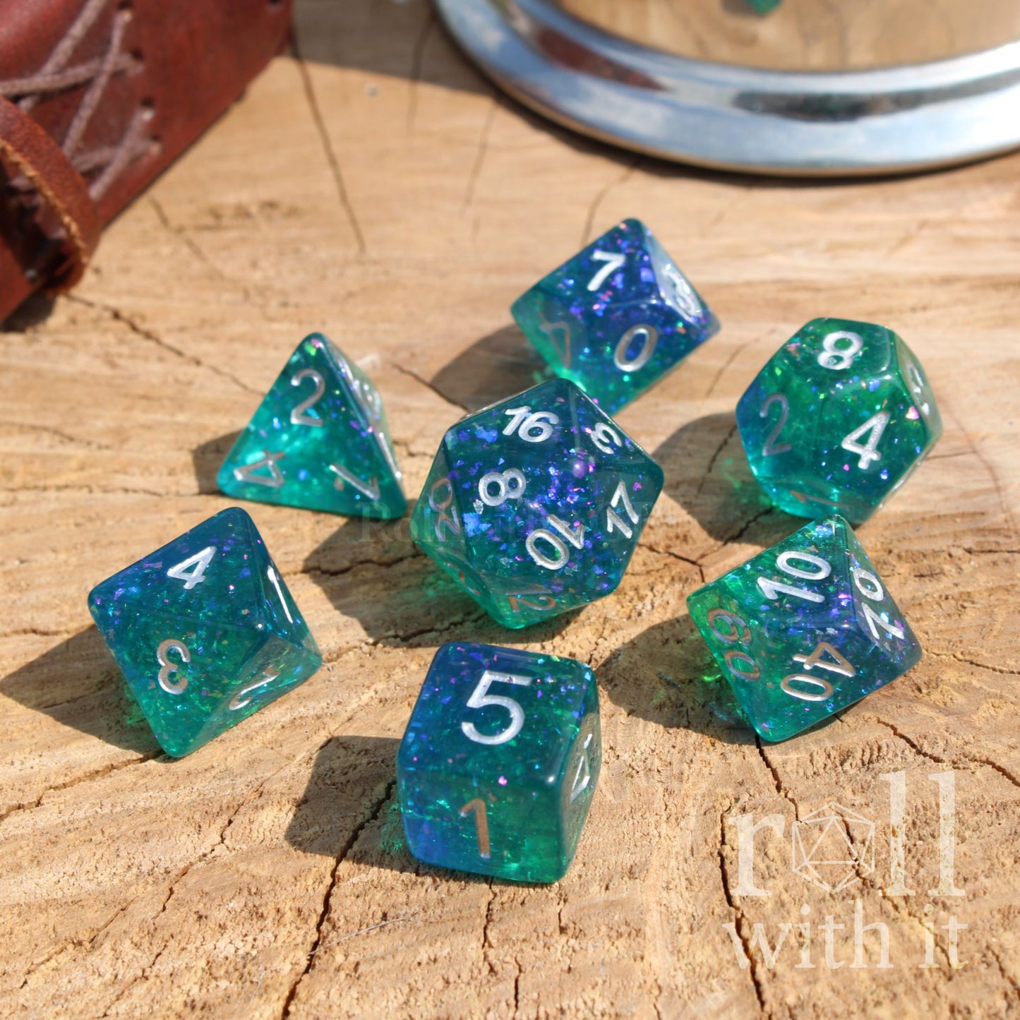 Set of teal reflective polyhedral dice with white numbers on a wooden surface, featuring the text 'roll with it'.