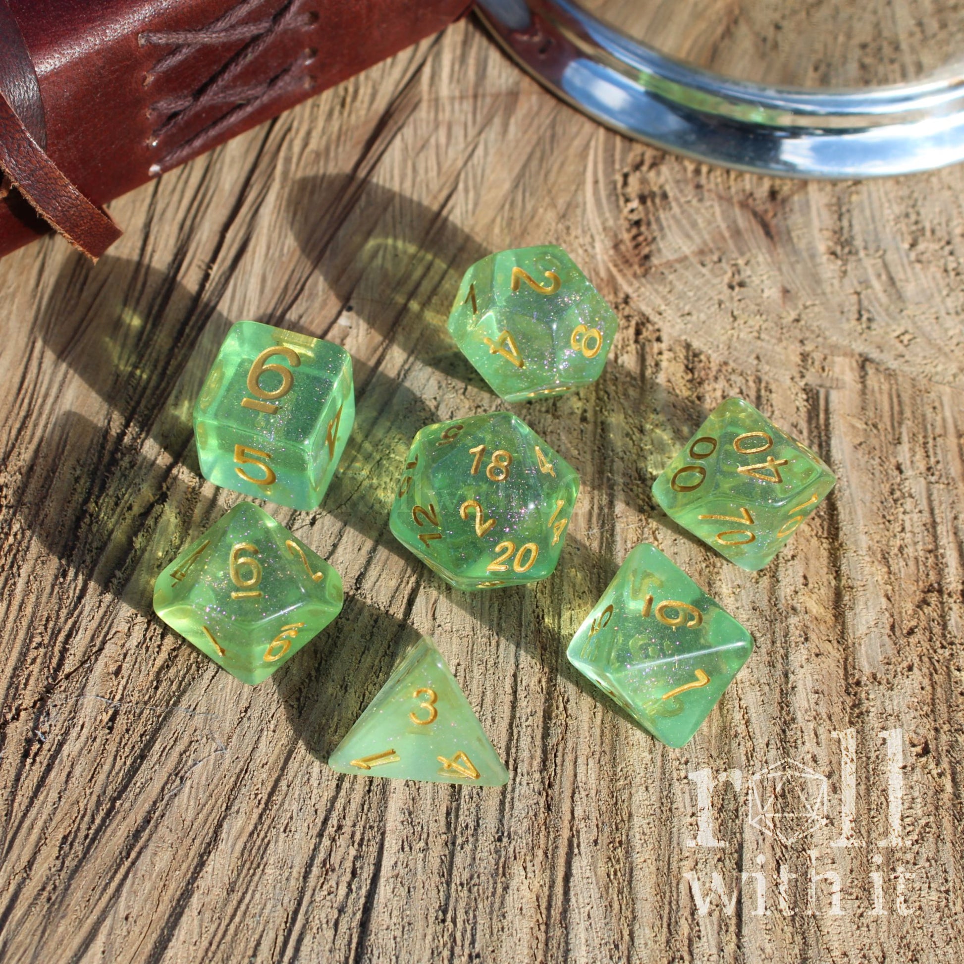 Set of green clear glittery shiny polyhedral dice with gold numbers on a wooden surface, featuring the brand 'Roll With It'.