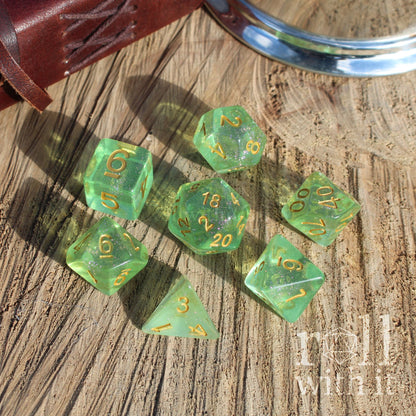 Set of green clear glittery shiny polyhedral dice with gold numbers on a wooden surface, featuring the brand 'Roll With It'.
