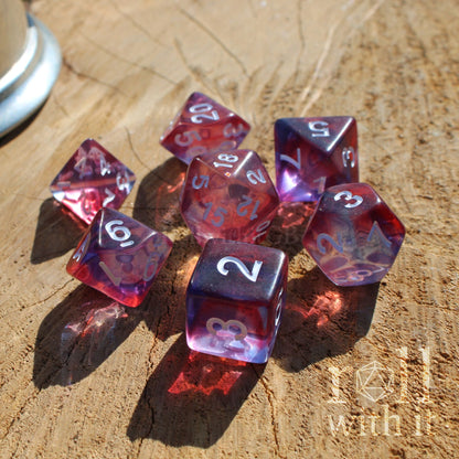 Set of red and purple mixed clear shiny polyhedral dice on a wooden surface with a brand logo in the corner.