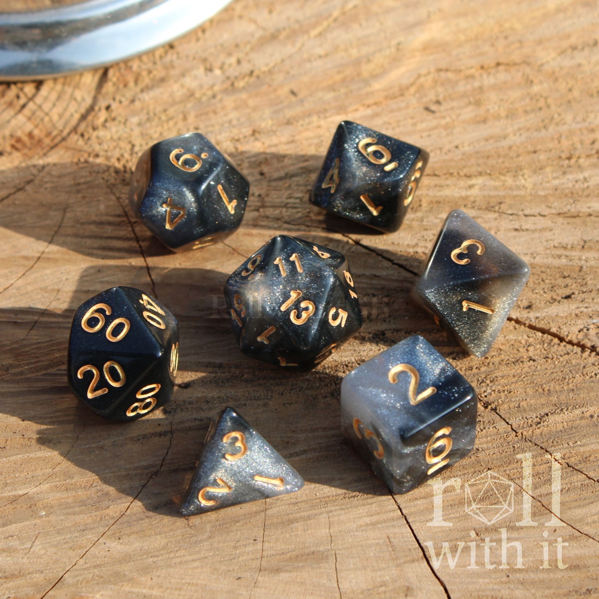 Set of metallic glitter coloured silvre polyhedral dice with gold numbers on a wooden surface, featuring 'roll with it' branding.
