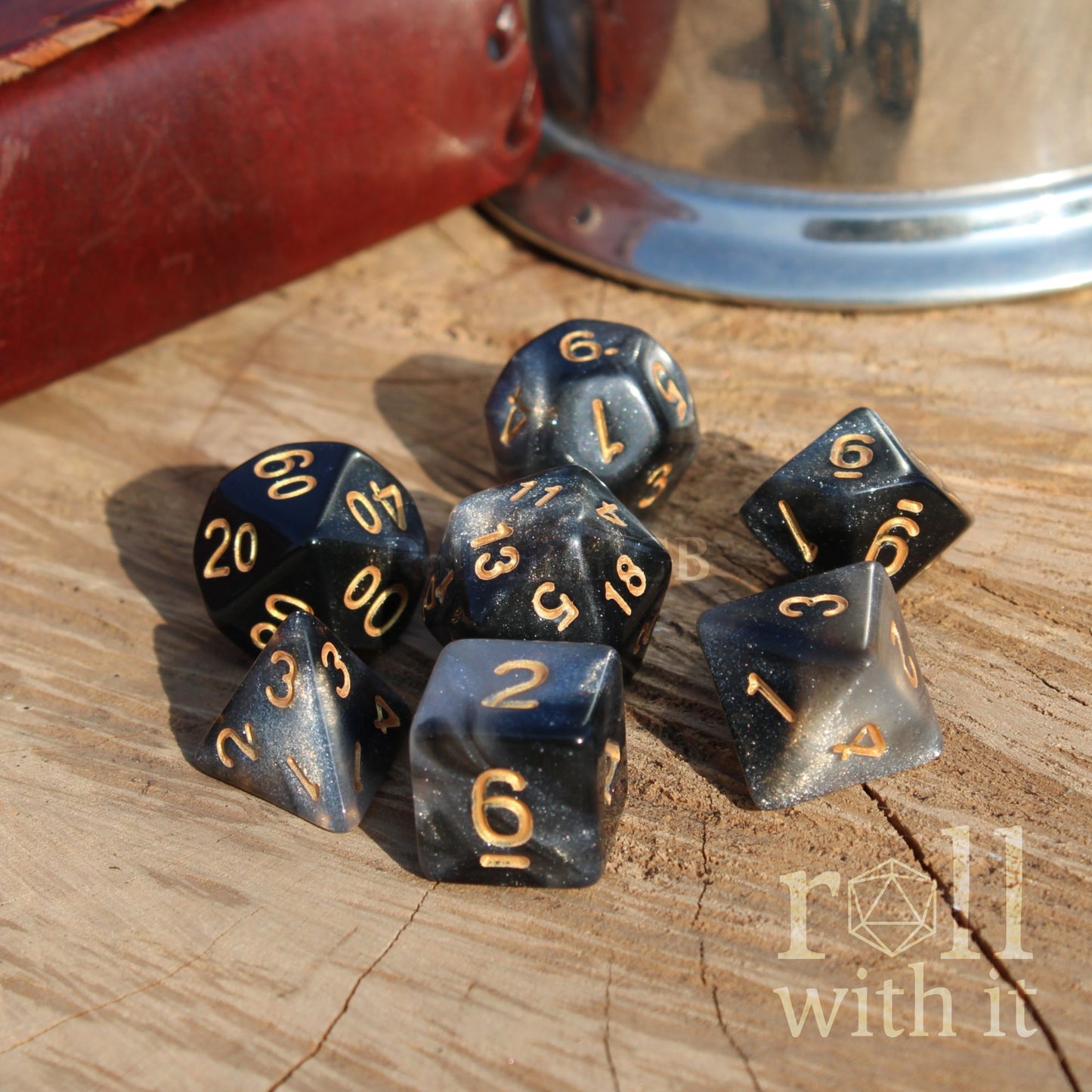 Set of black and silver star like polyhedral dice with gold numbers on a wooden surface, featuring 'roll with it' branding.