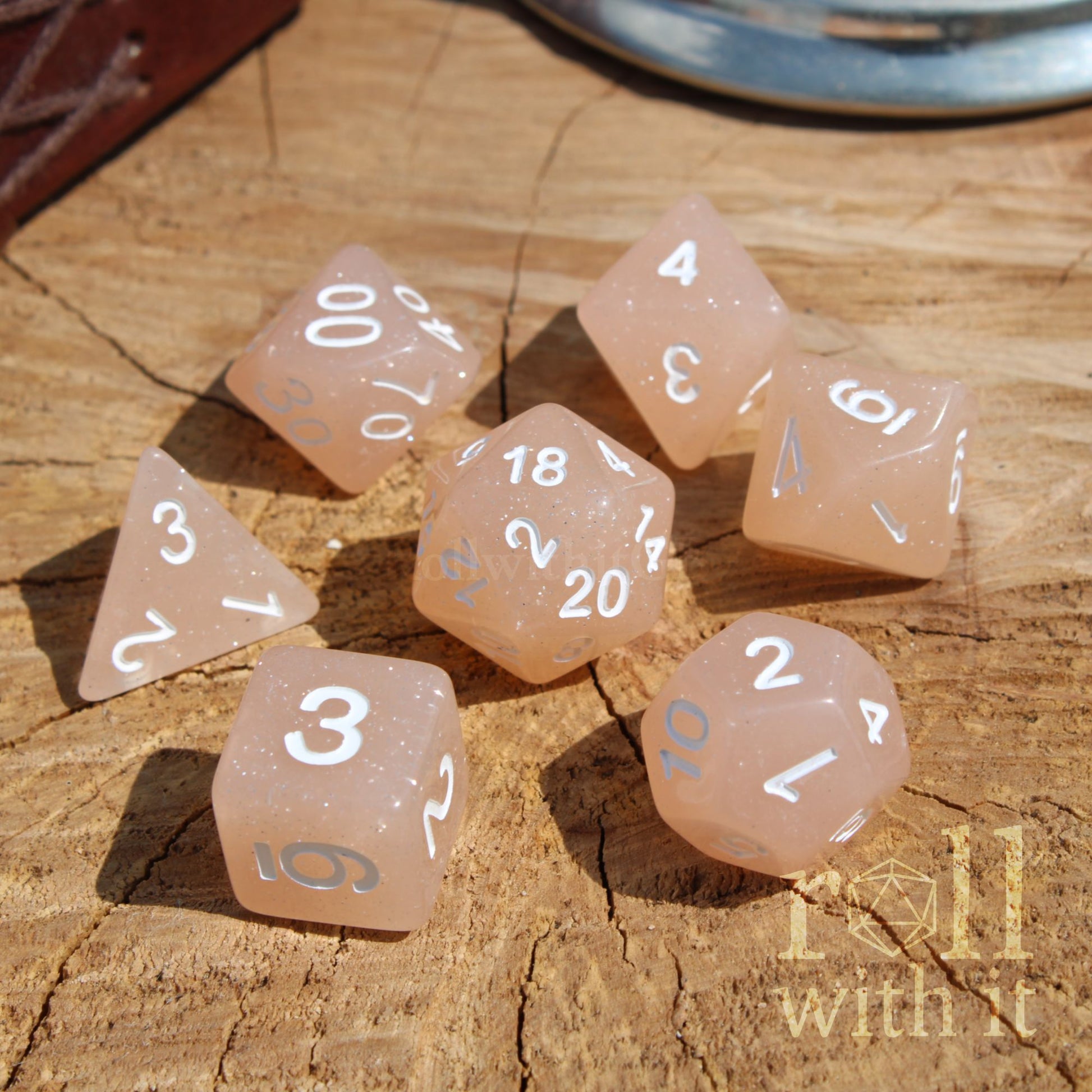 Set of simple glittery pink polyhedral dice with white numbers on a wooden surface, featuring the 'Roll with it' brand