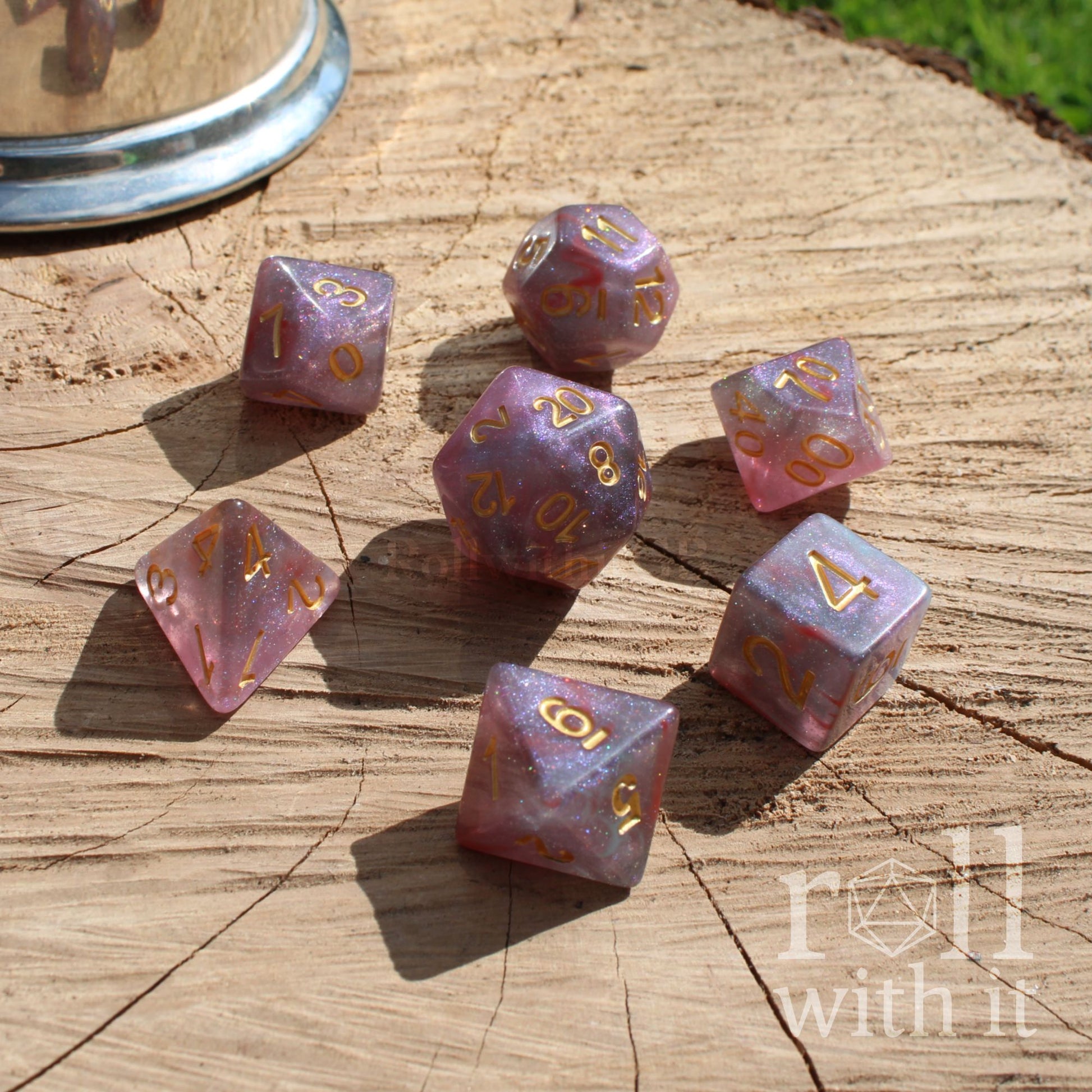 Set of glitter star like pink purple polyhedral dice with gold numbers on a wooden surface, featuring 'roll with it' branding.