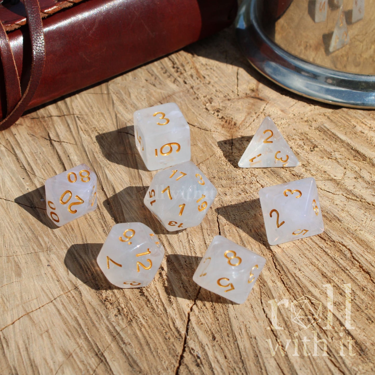 Set of white dice with gold numbers on a wooden surface, accompanied by a leather-bound book and a metal cup.