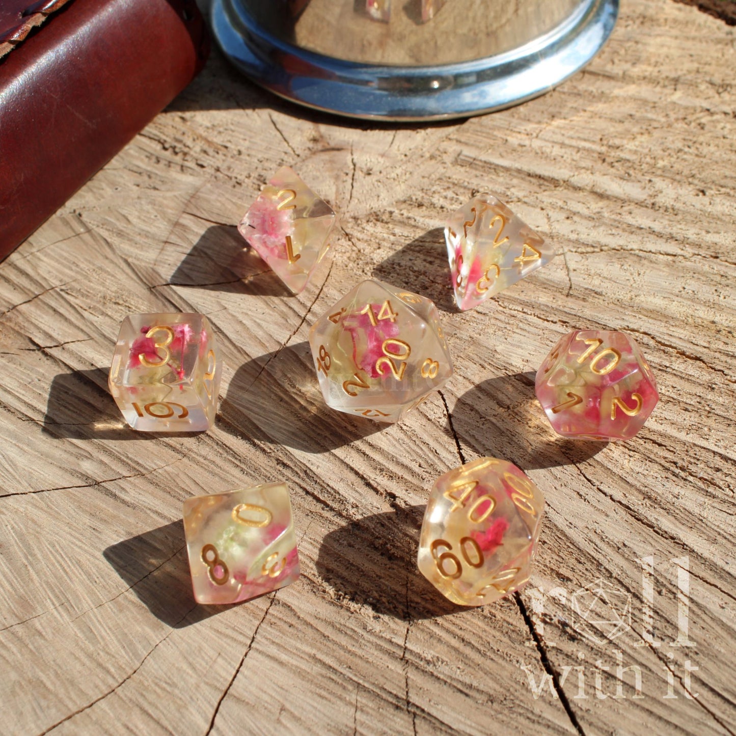 Set of 7 clear transparent dice with pink and gold miniature flowers and golden numbers on a wooden surface