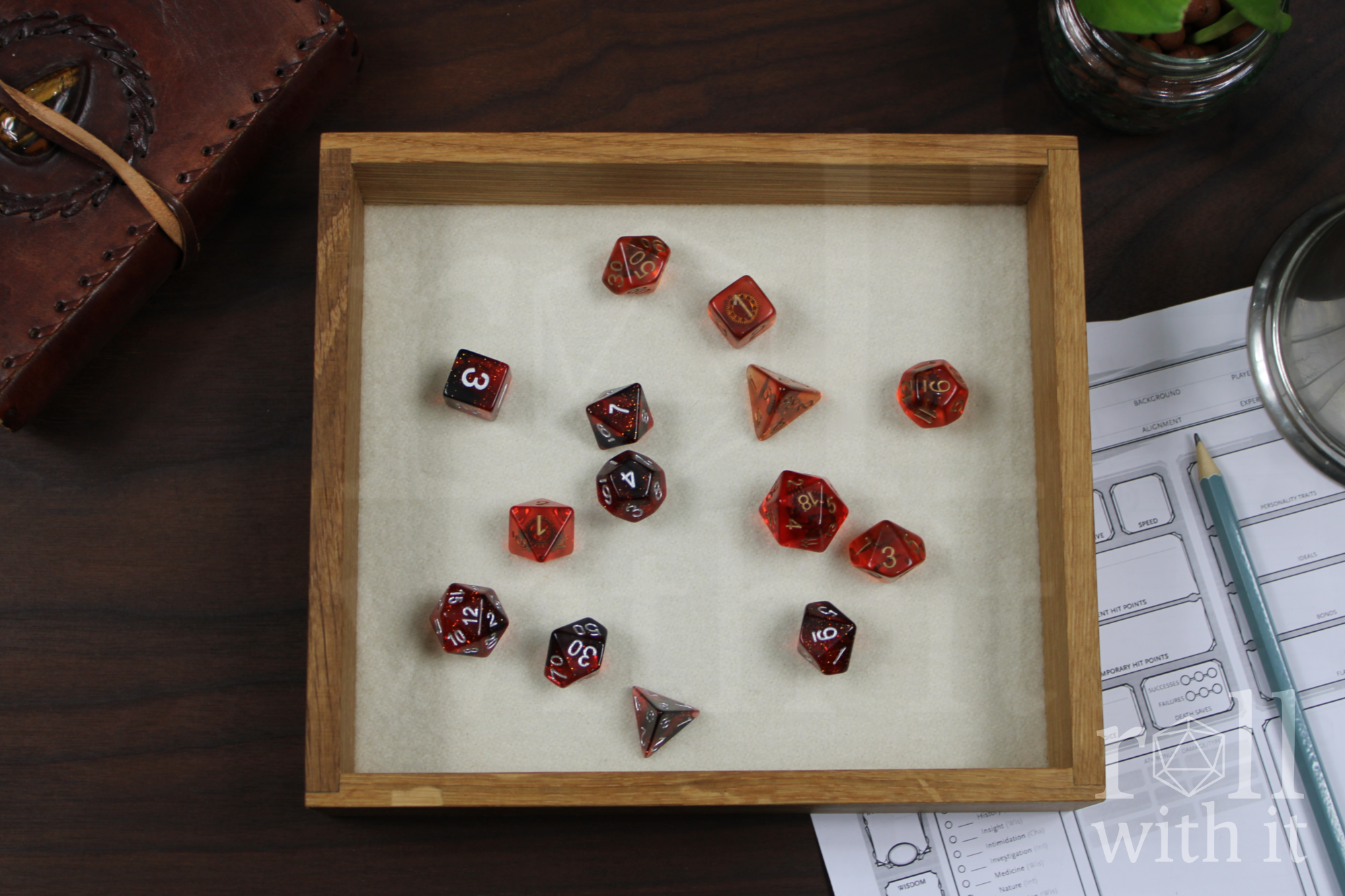 Orange, Red and black Dungeons and Dragons RPG dice sat on a cream white felt base in a wooden white oak tabletop dice tray on a table with 'roll with it' branding.