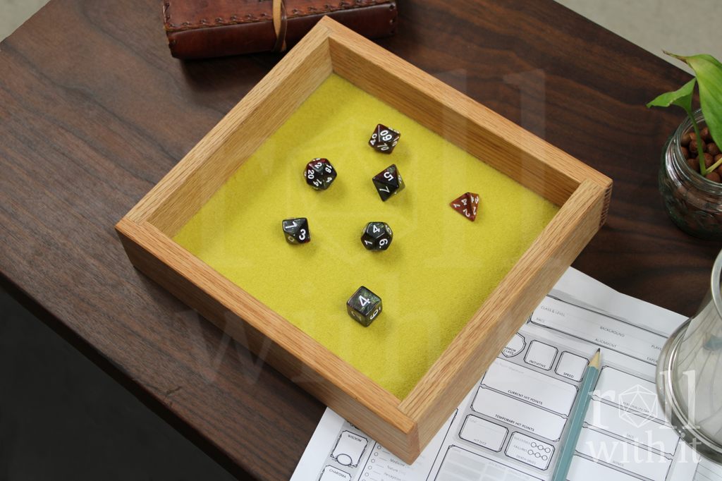 Wooden red oak dice tray with chartreuse yellow felt interior on a wooden table, featuring 'roll with it' logo. Red and black dice are inside the tray. Other tabletop roleplaying accessories are in the background, including a character sheet.