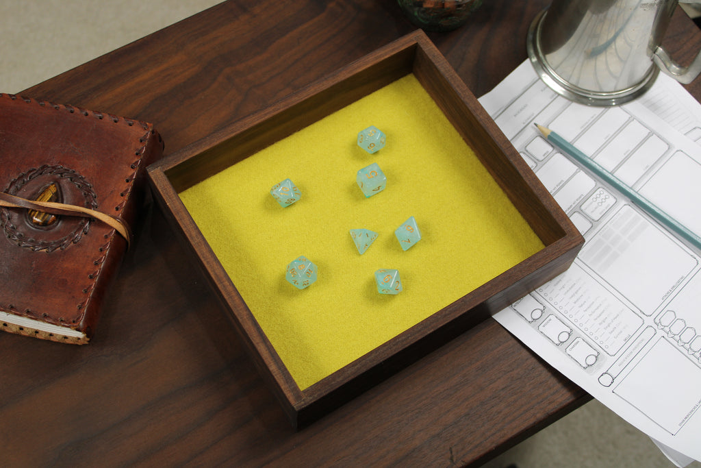 Solid walnut dice throwing tray with clear blue polyhedral roleplaying dice on a yellow felt base, next to a leather notebook and other dungeons and dragons accessories.