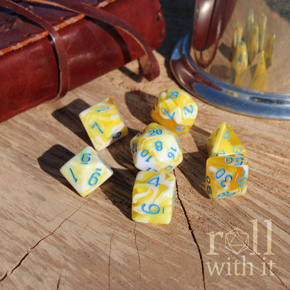 Yellow and white mixed marbled dice with blue numbers on a wooden surface, next to a leather-bound book and a metal cup.